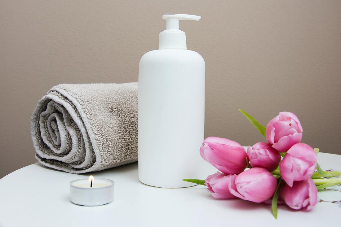 A picture of a tall, white bottle of lotiion on a small, white tabletop.  To the left of the lotion is a rolled up gray towel.  To the right are laying six pink tulips.  in front of the towel is a small votive candle.