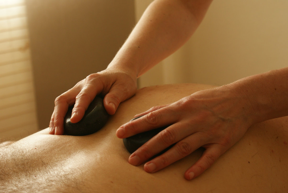 A picture of a man lying face down with a woman's hands rubbing two medium sized smooth, flat, dark stones on his back.