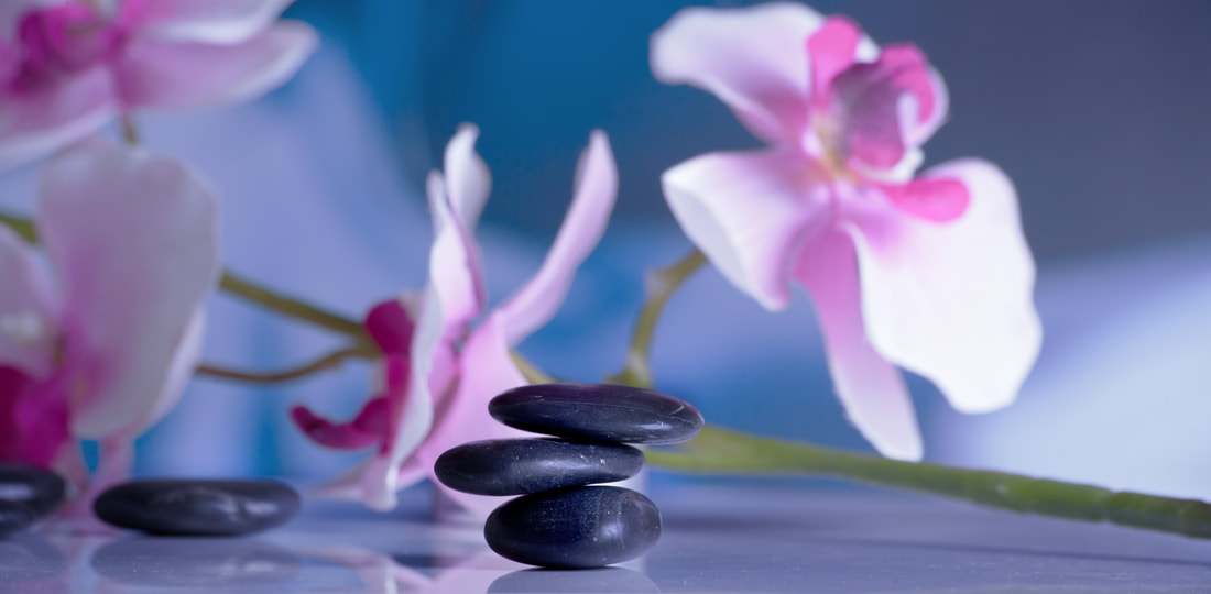 A picture of three small, dark, flat stones that are stacked.  To the left of the stacked stones are two more similar stones lying beside each other.  In the background are white and pink orchids.