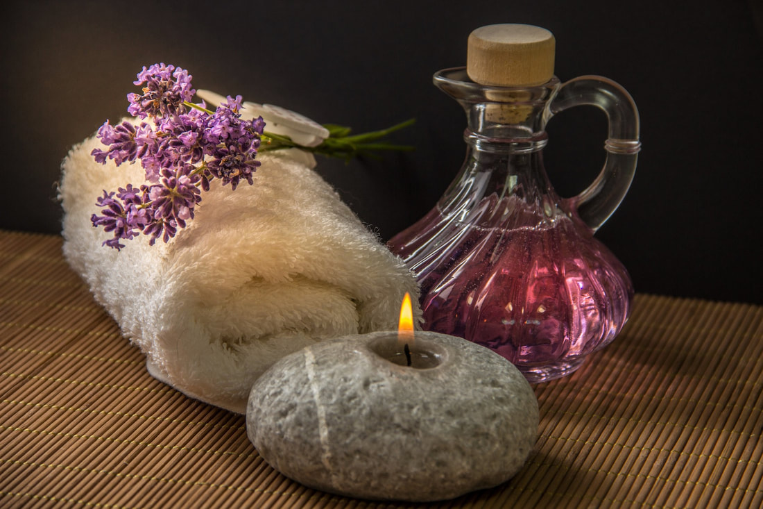 A picture of a rolled up white towel with some small, purple flowers lying on top.  To the right of the towel is an ornate glass jar with a handle and stopper.  Inside the jar is a pink, translucent liquid.  In front of the towel and jar is a lit candle that resembles a gray stone.