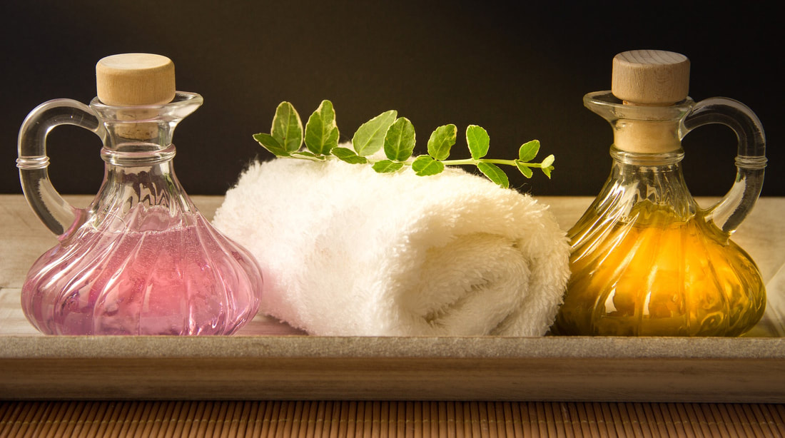A picture of a rolled up white towel on a tray with some sort of herb resting on top.  To the left of the towel is an ornate glass jar with handle and stopper.  Inside the jar is a pink, translucent liquid.  To the right side of the towel is an identical jar containing a gold, translucent liquid.