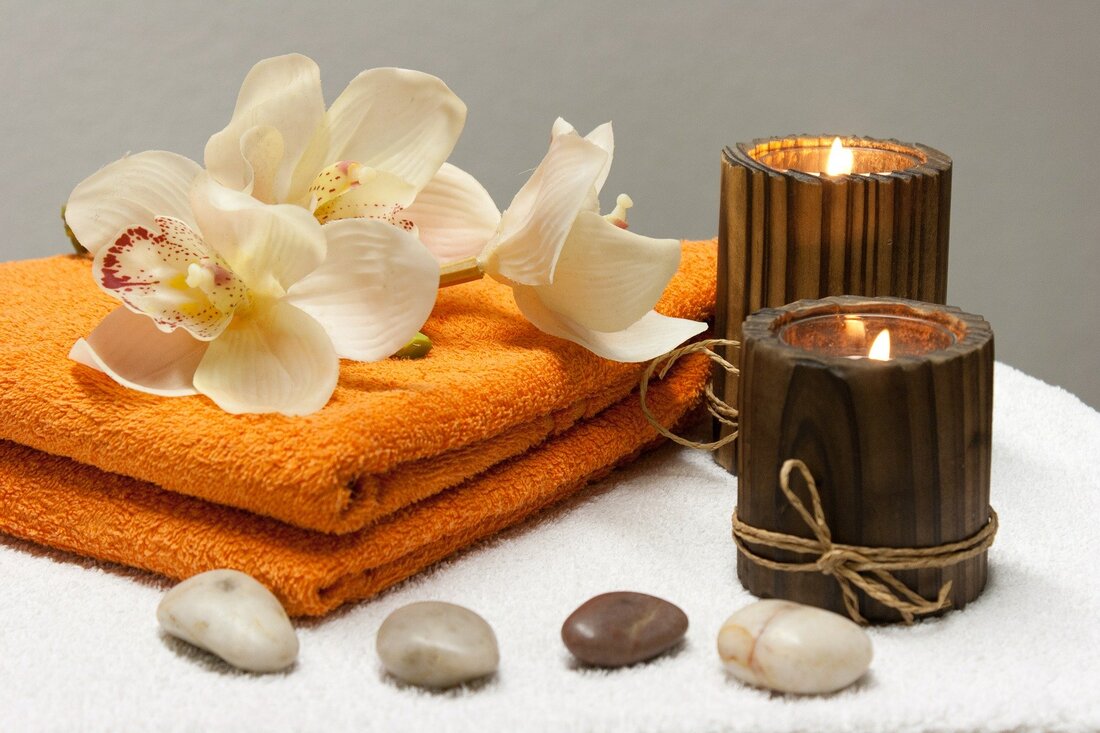 A picture of two lit, wooden candles on white terrycloth and two folded orange towels next to them.  There are three cream colored orchids on top of the towels and there are four small, smooth, alabaster and brown colored stones lined up in front of the candles and towels.